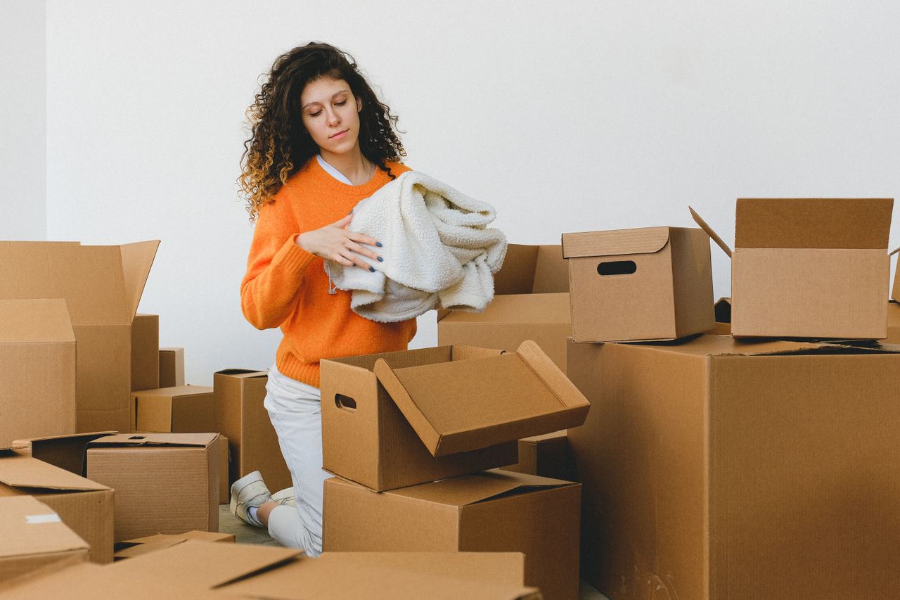 Concentrated young lady with curly hair standing on knees on floor and packing clothes into carton boxes before moving into new flat